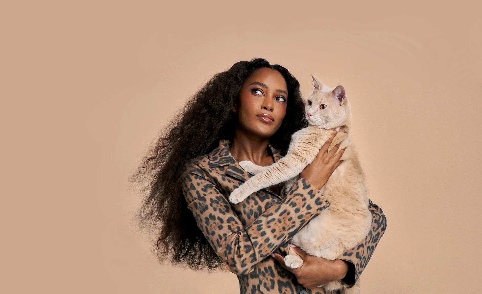 A woman in a leopard-print jacket holds a cream-colored cat against a tan backdrop